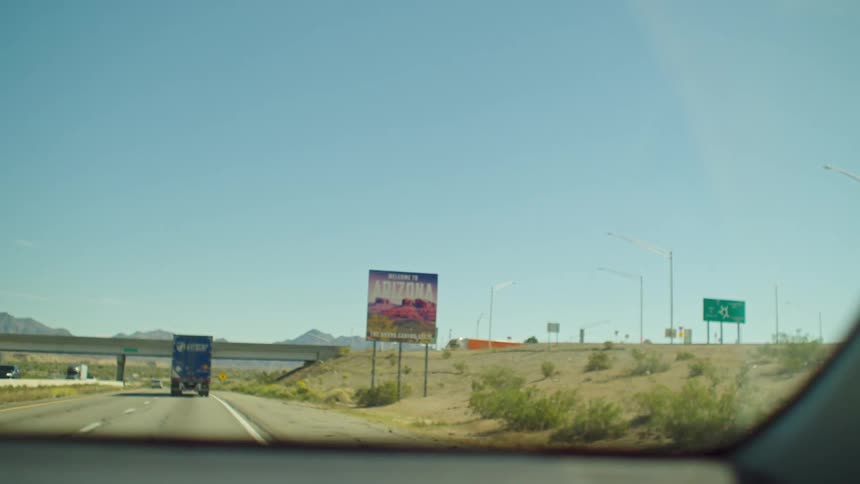 A point-of-view shot from a car approaching the 'Welcome to Arizona' sign on a desert highway on a sunny day