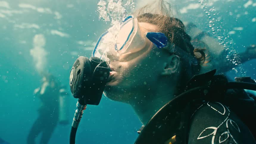 Close-up of a female scuba diver breathing underwater in slow motion as sunlight filters through the blue ocean