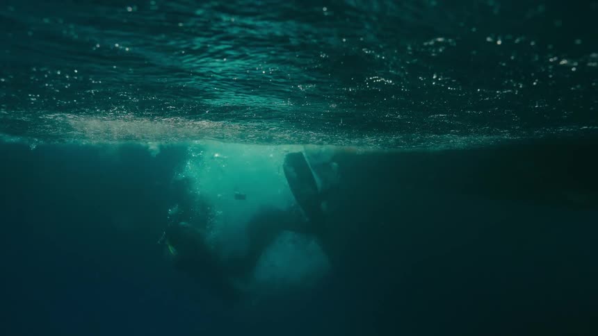 A female scuba diver performs a backward roll off a boat into the deep blue ocean, captured in a slow-motion over-under shot