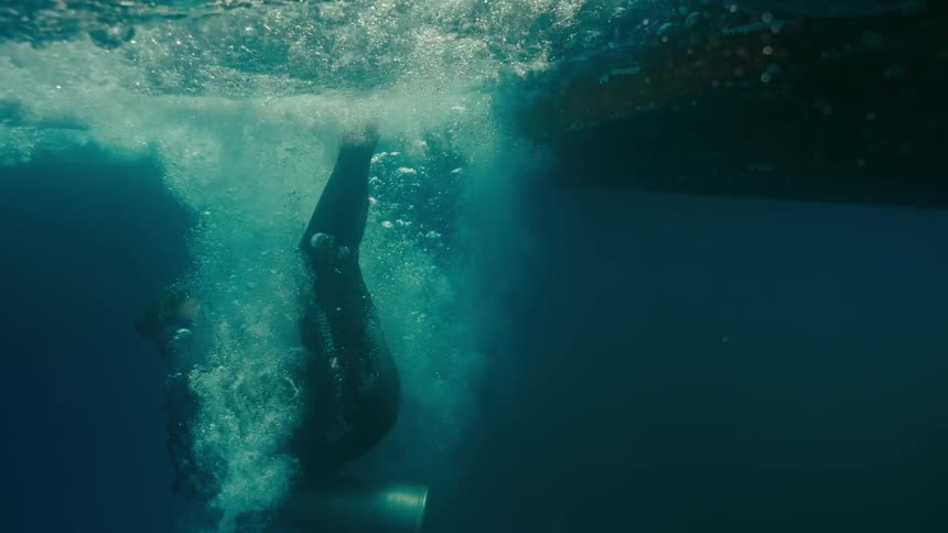 A scuba diver executes a slow-motion backward roll entry into the clear blue ocean, captured from a split-shot underwater perspective