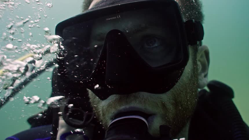 A super slow-motion, extreme close-up of a scuba diver's face as he breathes underwater, with bubbles rising from his regulator