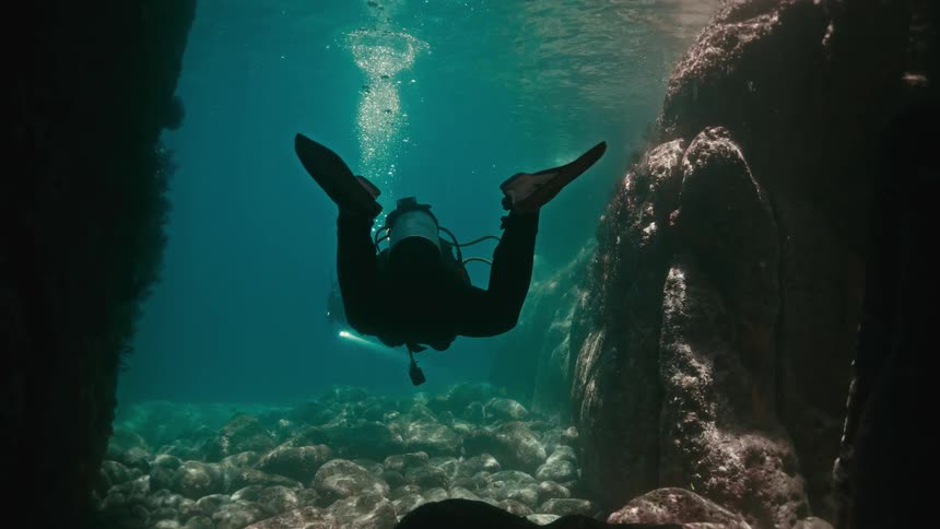 A silhouetted scuba diver floats peacefully in an underwater rock canyon as a second diver with a light emerges from the depths