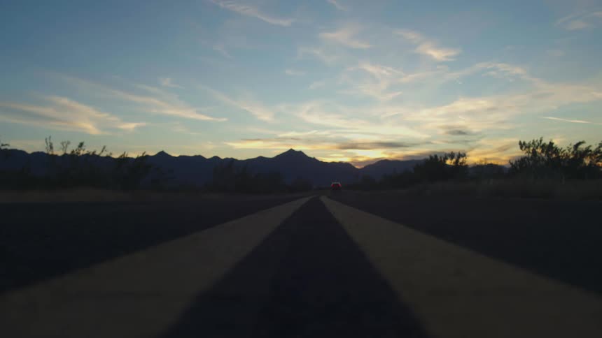 A low-angle shot captures an SUV driving down a desert highway towards a stunning mountain sunset