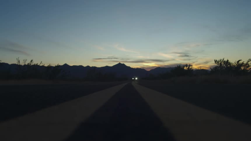 A lone SUV with headlights on drives toward and over the camera on a desolate desert highway at sunset