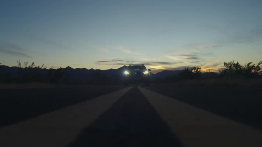 A lone SUV with headlights on drives toward and over the camera on a desolate desert highway at sunset