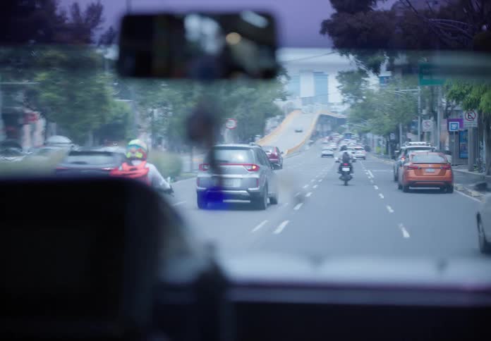 An interior point of view shot from a moving bus shows the city road ahead before focusing on protective charms hanging from the rearview mirror