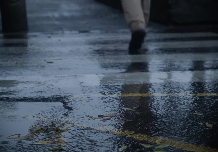 Raindrops fall onto a wet city crosswalk as a person in shoes and pants walks across in slow motion
