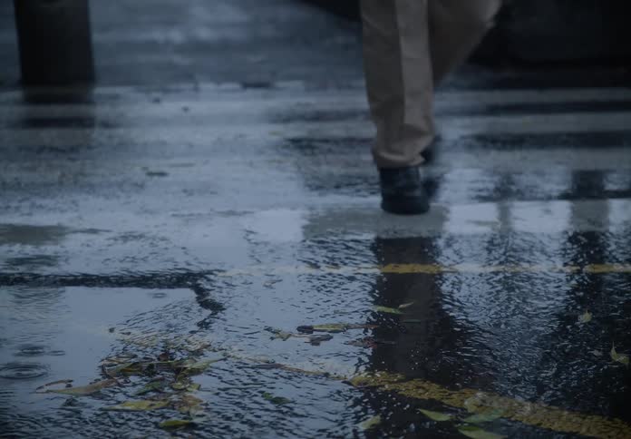 Raindrops fall on a wet city crosswalk as a person's feet and a car's tires pass by in slow motion