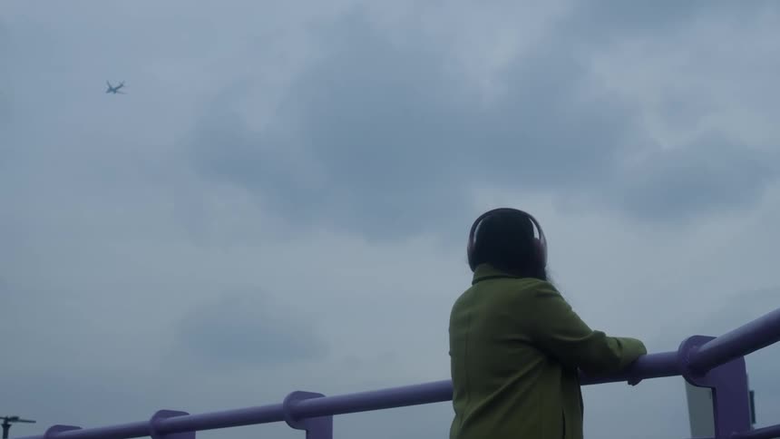 A lone person wearing headphones watches a distant airplane fly across a moody, overcast sky while leaning on a purple railing