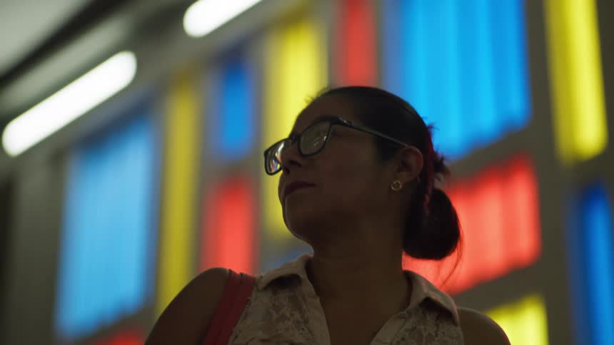 A pensive woman wearing glasses looks to the side against a brightly colored and abstract background of lights