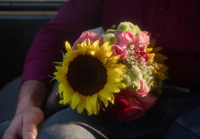 A man holds a vibrant bouquet of sunflowers and roses in his lap, waiting inside a car as the golden hour light shines in