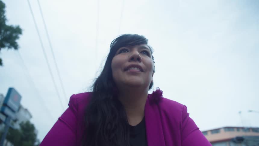 An empowered woman in a bold magenta blazer looks up with a hopeful smile while walking through the city streets in slow motion