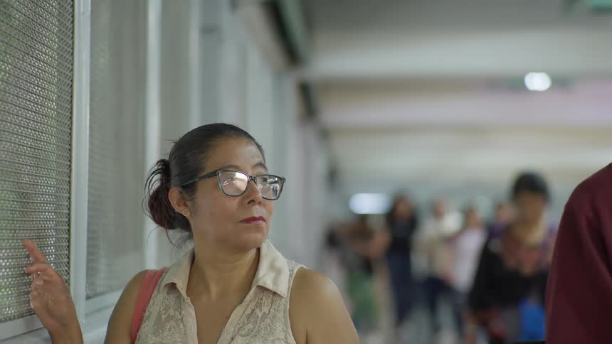 A Latin woman with glasses looks off thoughtfully while standing in a busy public transit hallway