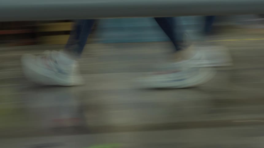 A low-angle, motion-blurred shot follows a person's feet in white sneakers as they walk quickly indoors