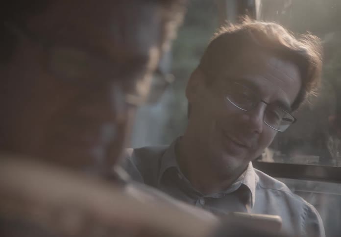 A smiling man wearing glasses uses his smartphone while commuting on a bus during a hazy golden hour