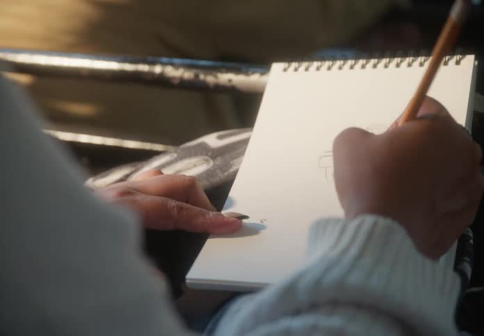 An over-the-shoulder shot captures a woman sketching in a notepad, then slowly reveals her face as she focuses on a screen while wearing headphones