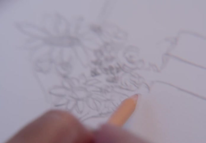 A soft focus, close-up shot of an artist's hand using a colored pencil to fill in a sketch of a flower bouquet