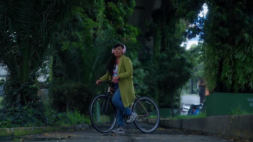 A woman wearing headphones leans on her bicycle while taking a break in a lush, green urban park under a highway overpass