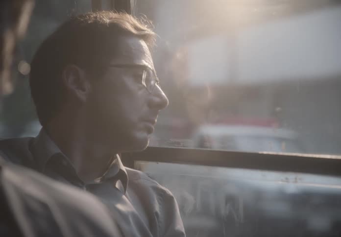 A contemplative man with glasses gazes out the window of a moving bus as the sunlight streams in
