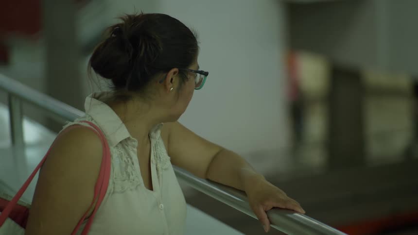 A woman with her hair in a bun stands waiting in a public space, holding onto a metal railing