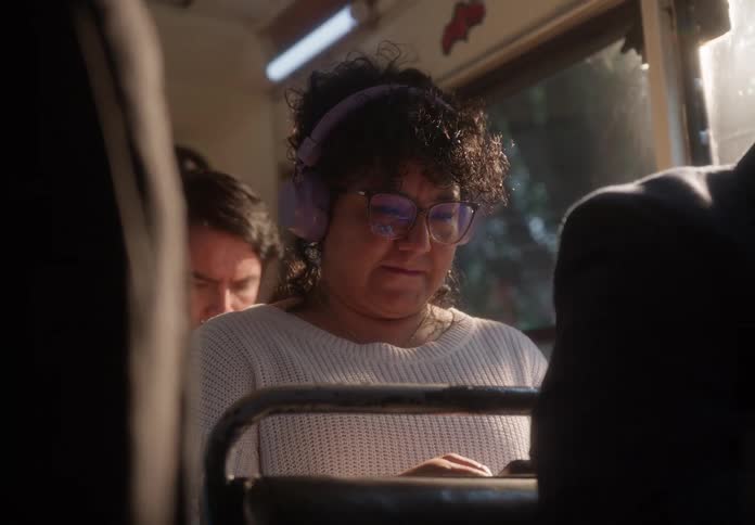 A woman with curly hair and headphones is focused on her phone while riding a bus during a sunny day