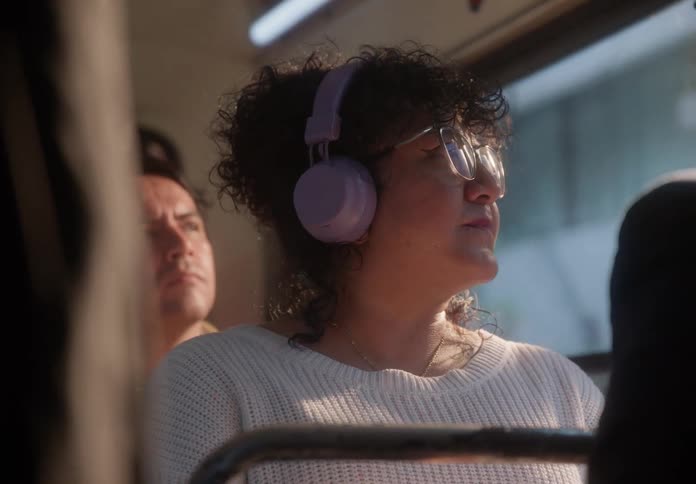 A woman with curly hair and headphones listens to music and peacefully enjoys the ride as warm sunlight streams through the bus window