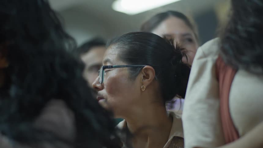 A pensive middle-aged woman with glasses stands in a crowd, her hair blowing gently in the breeze