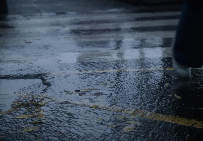 Slow motion view of a person walking across a rain-soaked street, their reflection visible on the wet asphalt