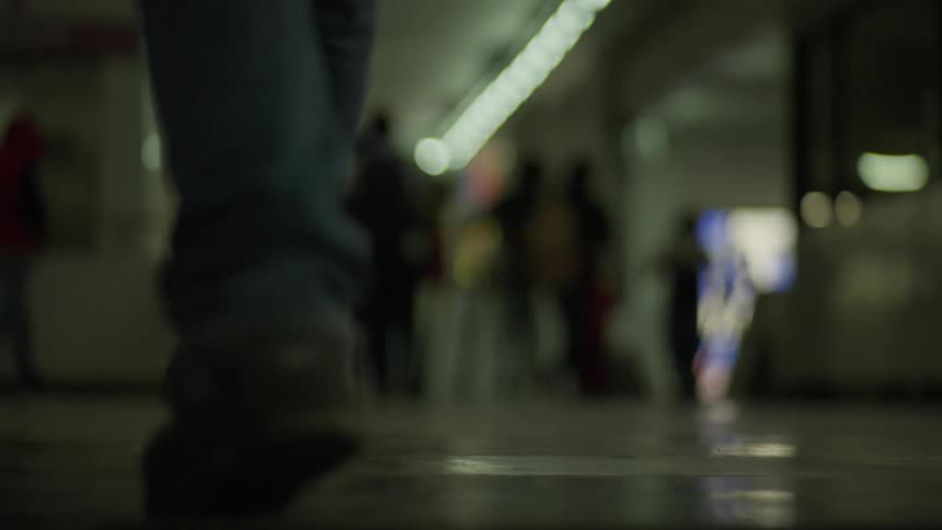 A slow-motion, low-angle, out-of-focus shot of a person walking away from the camera in a dimly lit underground station