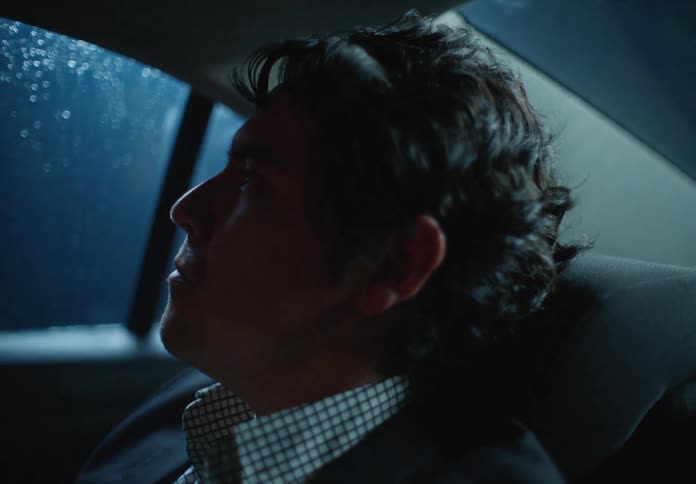A weary businessman dozes off in the backseat of a car during a rainy night commute through the city