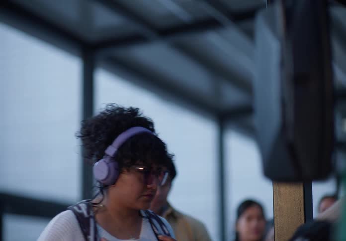 A woman with curly hair and headphones waits patiently in a line with other commuters at a transit station during twilight