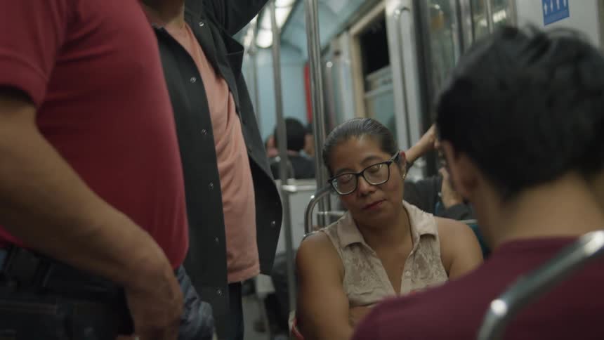 A tired woman wearing glasses dozes off while sitting amongst other passengers on a crowded subway car
