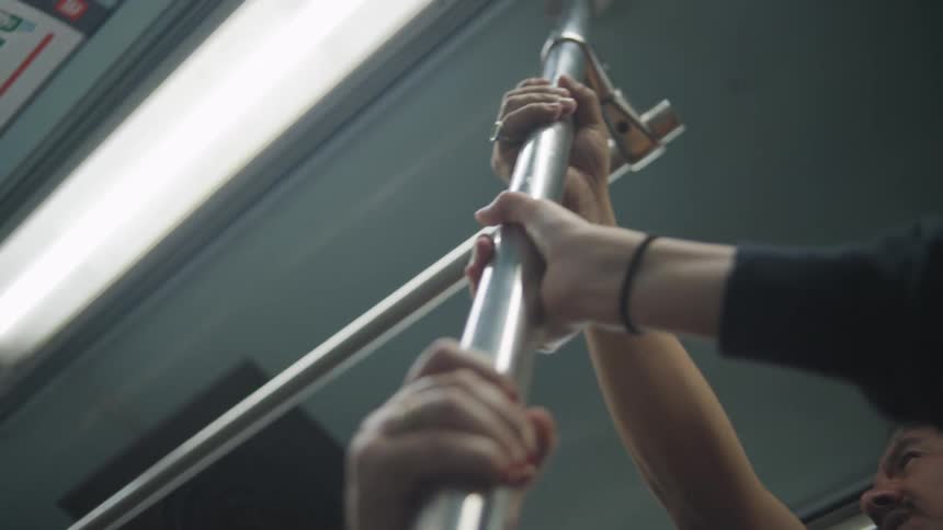 A low-angle close-up captures multiple passengers' hands holding onto a metal pole for balance on a moving subway train