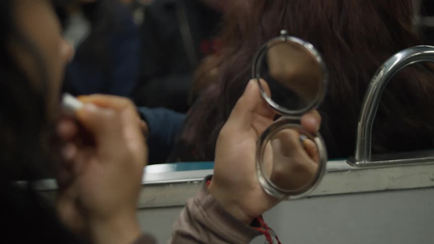Over the shoulder shot of a woman applying lipstick in a compact mirror while riding on a crowded train