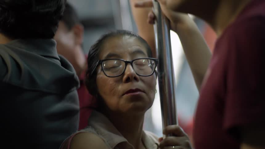 A tired woman with glasses holds a pole and closes her eyes on a crowded, swaying subway train