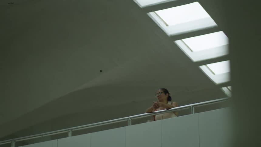 A woman with glasses stands pensively on an upper-level walkway inside a vast, modern architectural space with a curved ceiling