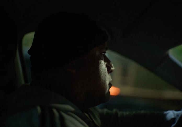 A serious man with neck tattoos drives his car through the city streets at night, his face lit by the passing lights