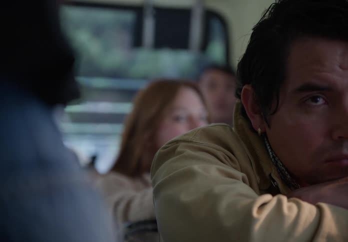 A tired man in a tan jacket wakes up and looks up with a pensive expression while riding on a bus