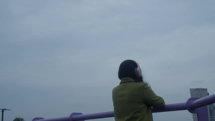A woman seen from behind puts on headphones and leans against a railing to enjoy a contemplative moment on a gloomy, overcast day