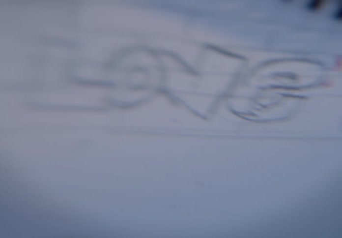 A soft-focus close-up shows a hand sketching the word 'LOVE' in a notebook before erasing other handwritten text on the page