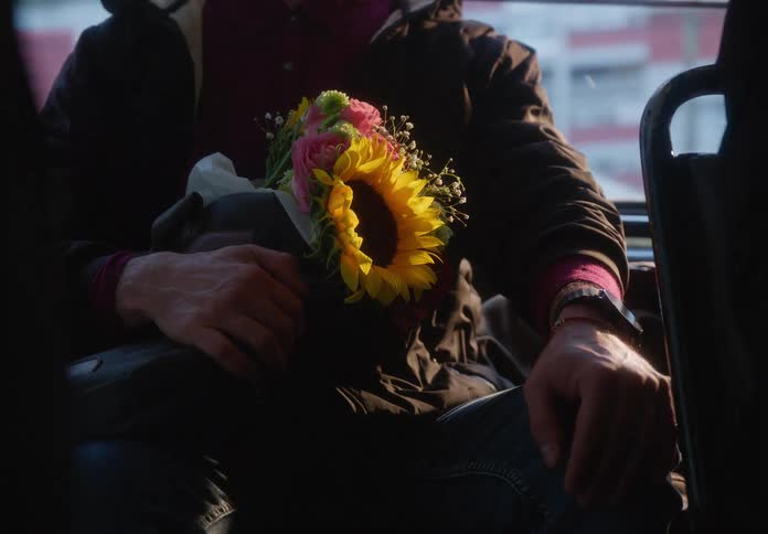 Sunlight illuminates a man holding a bright bouquet of sunflowers and roses while riding on a city bus