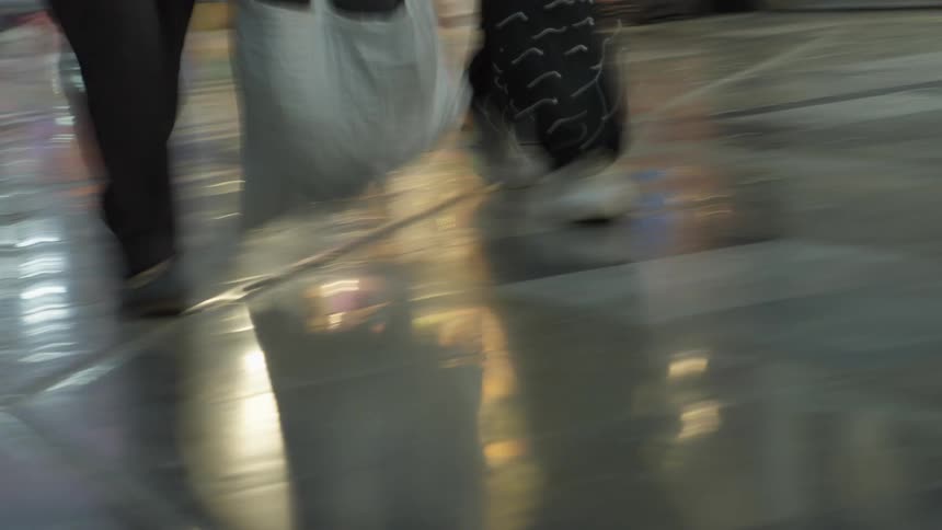 An abstract, low-angle, motion-blur shot shows people's legs walking across a shiny, reflective floor in a bustling public space