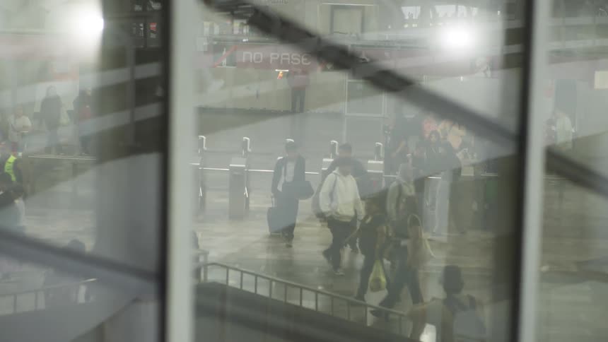 Slow motion footage shows commuters walking through a busy transit station as seen through a reflective glass window