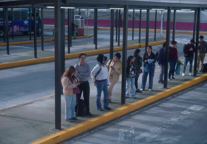 A diverse group of commuters waits patiently at a bus terminal before a minibus arrives to pick them up