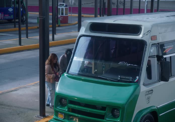 A diverse group of commuters waits patiently at a bus terminal before a minibus arrives to pick them up