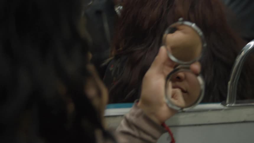 A woman applies lipstick in a compact mirror during her daily commute on a crowded subway train