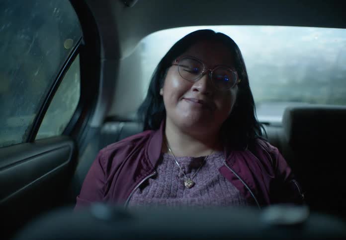 A woman sitting in the back of a car at night looks out the window thoughtfully before turning and smiling