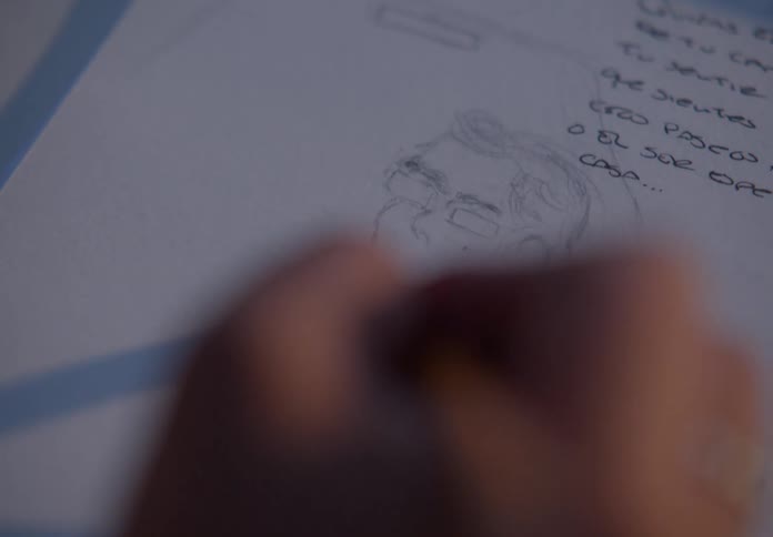 An intimate, soft-focus close-up of an artist's hand sketching a portrait of a person with glasses