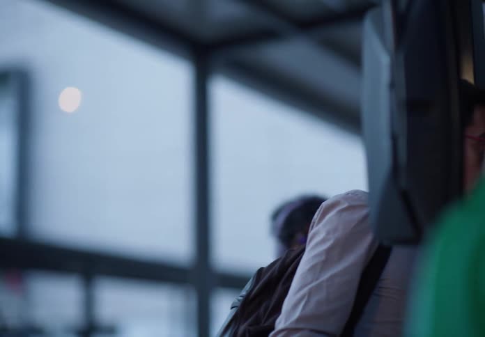 A weary middle-aged man wearing glasses waits for public transport and then puts on his backpack to board with other commuters