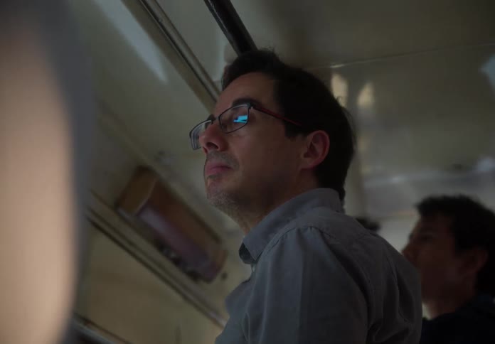 A pensive man with glasses holds onto a bar while standing on a crowded bus during his commute